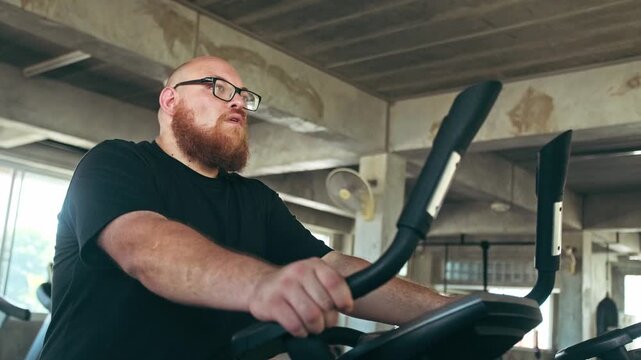 Bearded person using a stationary bike in an industrial-style gym, focused on an indoor cardio workout.