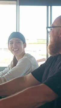 Two people chat and smile while pedaling on exercise bikes in a bright gym, enjoying a cardio workout beside large windows.