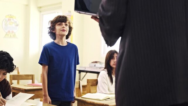 A boy stands beside his desk in a bright classroom, looking up at a teacher holding a device, while classmates work at their seats.
