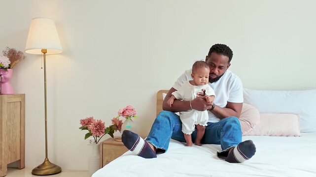 Smiling man on a bed holds a baby in a bright, cozy bedroom with flowers and a floor lamp, capturing a warm, playful bonding moment at home.