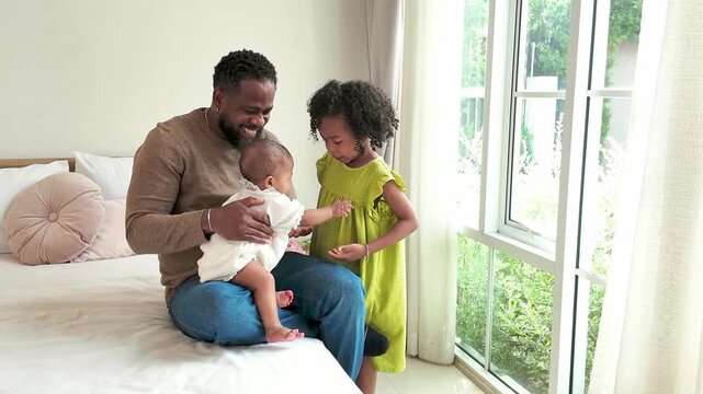 A father sits on a bed by a bright window, holding a baby while an older child smiles beside him. A cozy, warm family moment at home.