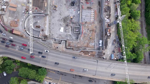 Drone video captures a straight-down aerial view of a massive construction site with two tower cranes, excavators, active machinery, and busy urban roads.