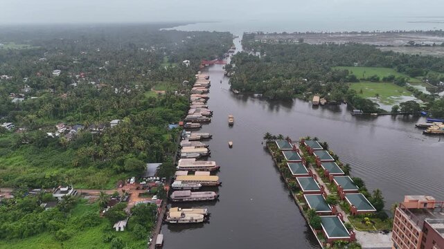 High-altitude aerial drone shot featuring a wide backwater canal in Alappuzha with traditional houseboats docked along the shoreline, waterfront resort cottages, and lush coconut palm groves.
