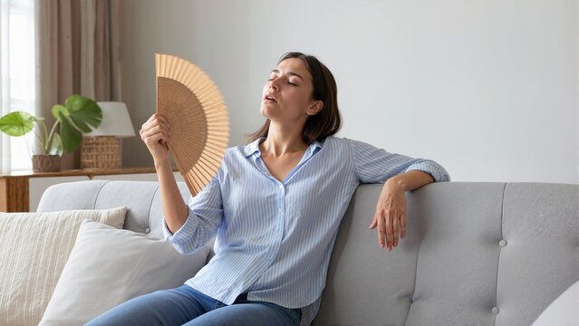 Woman cooling off with a wooden hand fan on a hot summer day. Exhausted female sitting on a sofa at home during a heatwave. Indoor temperature and climate control concept
