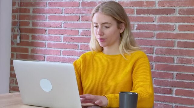 Beautiful young blonde woman in a yellow sweater finishing her work, closing the laptop, and getting up from her desk in a modern loft office with a red brick wall background