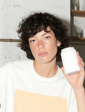 Young woman in light t-shirt holding aluminum soda can and looking directly into camera lens against low-key lighting background. Aluminum container with fizzy carbonated water.
