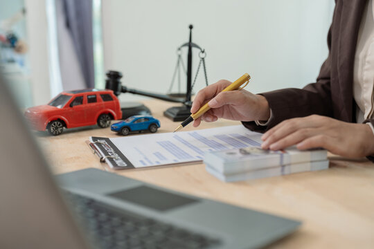 Lawyer or insurance agent holding pen over car loan contract agreement with toy cars, gavel, scale of justice and banknotes on desk