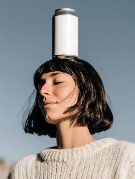 Young woman in woolen sweater with aluminum soda can on her head, with closed eyes and peaceful expression. Tin can of soda for food industry product catalog filling.