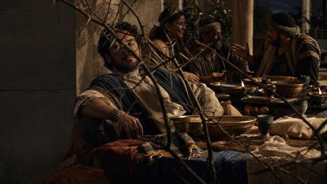 Man sitting at table with group of people in ancient clothes. Scene depicting biblical parable of sower illustrating distraction by worldly wealth and life pleasure. Faith concept.