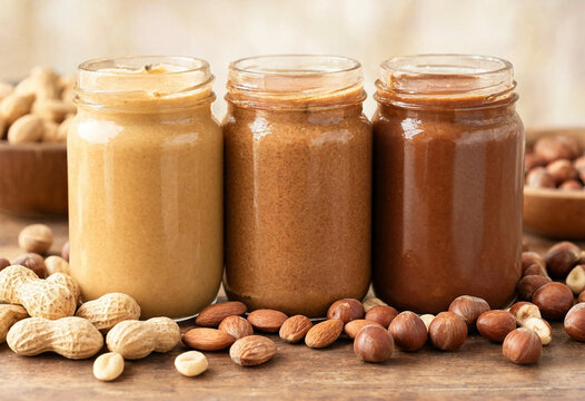 Three jars display peanut, almond, and hazelnut butter in different tones on a rustic wooden surface with scattered nuts. The arrangement highlights texture and natural ingredients for food packaging