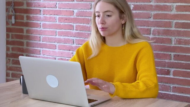 Young tired student working on a laptop, feeling bored and unmotivated while studying or working from home, she's drinking coffee to stay awake in front of a brick wall