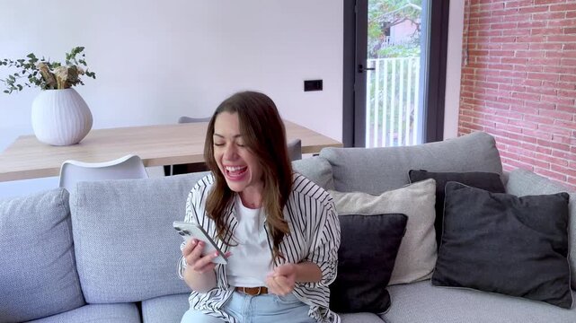 Happy young woman sitting on the couch at home, celebrating a victory or receiving good news on her smartphone with a fist pump. Overjoyed emotions after a successful outcome or achievement