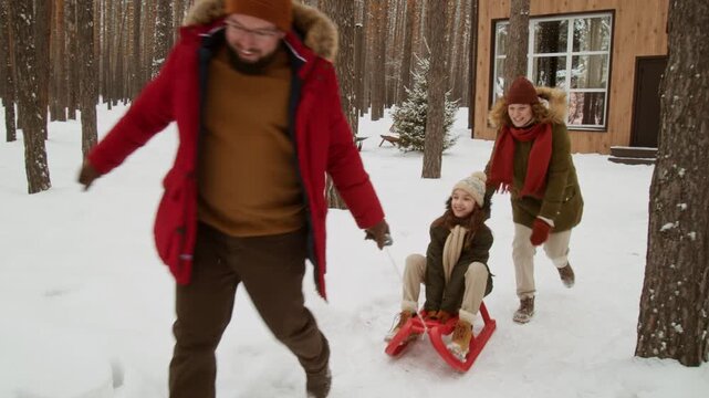 Caucasian middle aged parents pulling and pushing young daughter on sled in snowy pine forest near modern cabin. Footage is ideal for winter tourism campaigns and family holiday promotions
