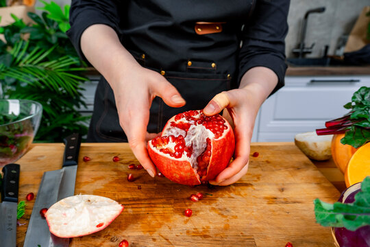 Woman chef breaks fresh red pomegranate on wooden cutting board in kitchen. Concept of healthy vegetarian diet and culinary preparation of organic food.