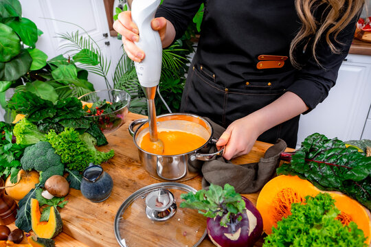 Nutritionist cook blends orange pumpkin vegetable soup with white immersion blender on wooden table. Organic vegetarian diet and culinary process with fresh seasonal ingredients.