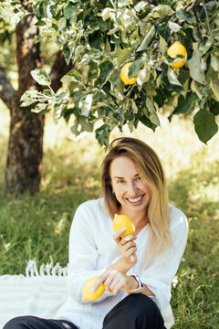 Smiling woman holding fresh lemons while sitting under a lemon tree in a sunny orchard
