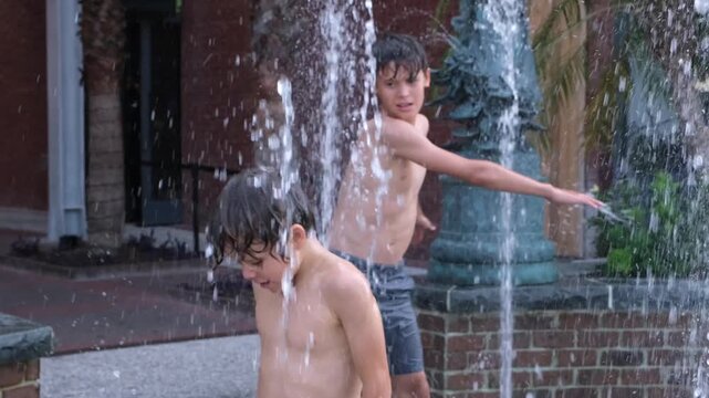 Two boys enjoy cooling off beneath the water jets of an outdoor splash fountain in a residential setting. The candid scene captures carefree childhood, active play.