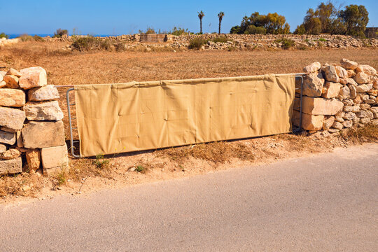 Burlap covered gate spans a gap in traditional stone wall along a rural Mediterranean road, with arid fields and palm trees beyond