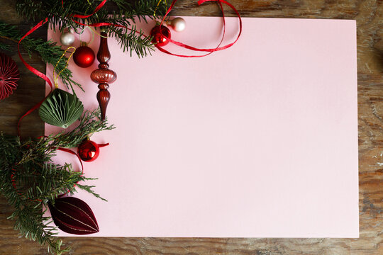 Elegant Christmas flat lay frame with paper ornaments, red baubles, glass finial, and fir branches on pastel pink background. Xmas background with copy space. Merry Christmas