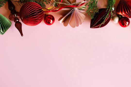 Stylish Christmas top border with paper ornaments, glass finial, red baubles, and fir branches on pastel pink background. Xmas flat lay woth copy space