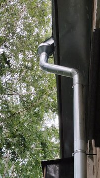 Rainwater flows down a metal drainpipe from a black roof of an old brick building. Green birch trees sway in the strong wind during a rainy summer day. Weather and outdoor concept.