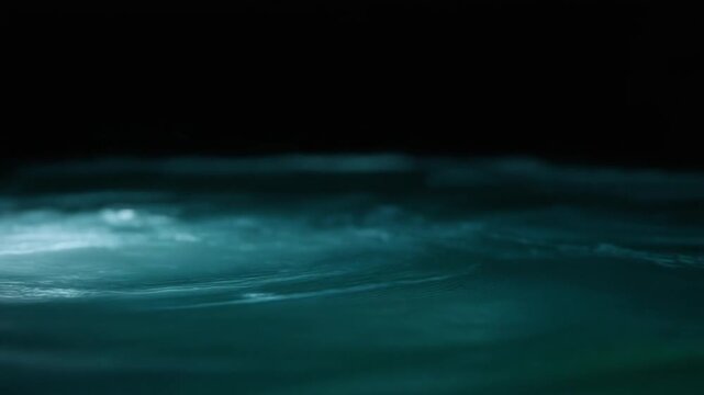 Dark teal-blue water swirls in continuous gentle currents, soft light revealing moving surface texture and subtle depth against near-black background with tranquil natural atmosphere.
