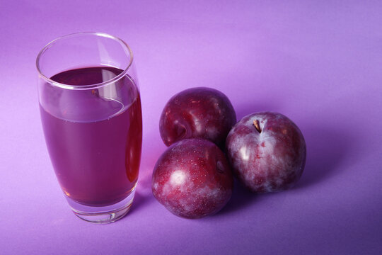 Vaso de zumo de ciruela natural acompa&ntilde;ado de ciruelas frescas sobre un fondo morado. Concepto de alimentaci&oacute;n saludable, bebida natural, fruta fresca y fotograf&iacute;a de producto en estudio.