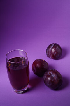 Vaso de zumo de ciruela natural acompa&ntilde;ado de ciruelas frescas sobre un fondo morado. Concepto de alimentaci&oacute;n saludable, bebida natural, fruta fresca y fotograf&iacute;a de producto en estudio.