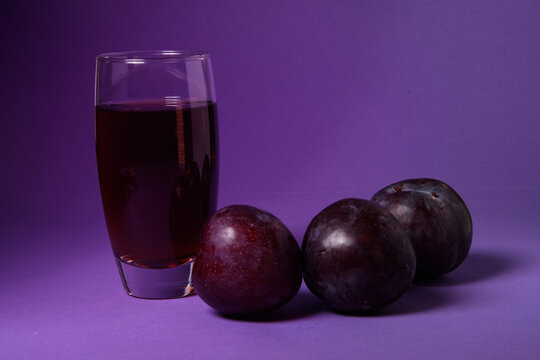 Vaso de zumo de ciruela natural acompa&ntilde;ado de ciruelas frescas sobre un fondo morado. Concepto de alimentaci&oacute;n saludable, bebida natural, fruta fresca y fotograf&iacute;a de producto en estudio.