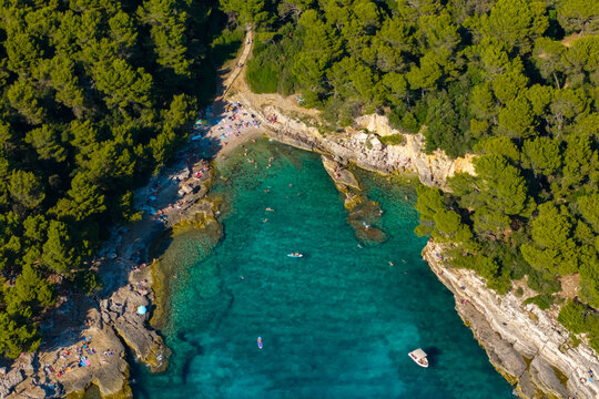 Top-down aerial view of crystal-clear turquoise water, natural sea caves, limestone cliffs, and boats anchored in the protected cove near Pula, Croatia