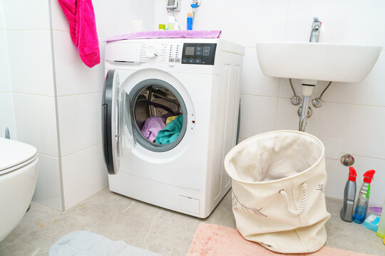 Modern front-loading washing machine with colorful clothes inside, positioned in a tiled bathroom beside a laundry basket and cleaning supplies. Domestic organization, laundry care, cleanliness