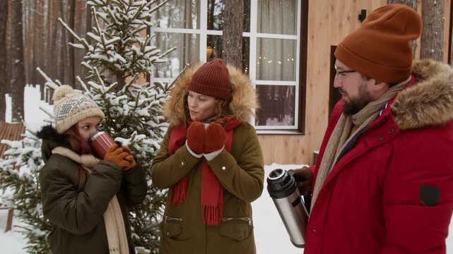 Caucasian family standing outdoors in winter, drinking hot beverages from thermos near wooden cabin. Perfect for winter tourism, family holiday promotions, and outdoor gear advertisements