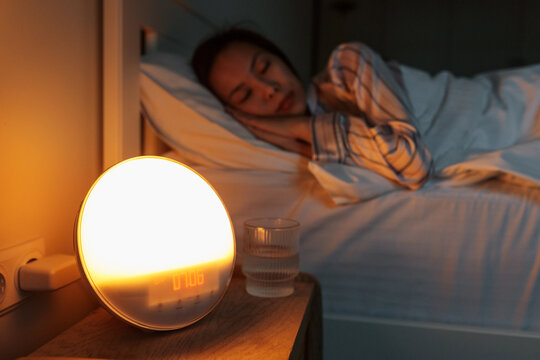 Selective focus on sunrise simulation artificial dawn alarm clock standing on bedside table with girl nicely sleeping in soft bed on background for circadian rhythm regulation and gentle waking-up