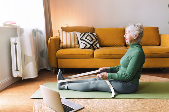 Senior woman training at home, sitting on green mat in fitness clothes and using rubber resistance band, following online video tutorial on laptop, doing physical exercise for strong muscles and bones