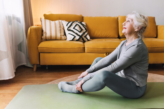 Aged fitness woman with gray hair enjoying her morning workout, practicing yoga at home, sitting in leggings on mat in butterfly position and looking at window, yellow sofa on background