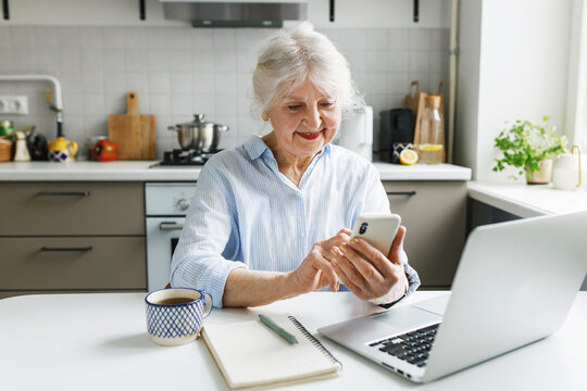 Portrait of curious elderly lady exploring Ai and prompt engineering, smiling at smartphone while messaging chat bot sitting at kitchen table with laptop and cup of tea. Digital literacy for seniors