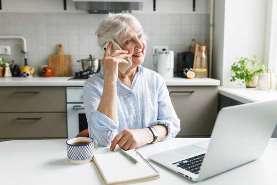 Senior female customer calling to clients support office to leave positive feedback on purchase or received services, sitting at kitchen table smiling while talking on phone and looking aside