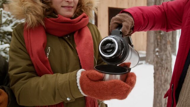 Young Caucasian woman holding metal cup while man pours hot tea from thermos in snowy forest. Footage is perfect for winter tourism, outdoor recreation, and travel gear marketing campaigns