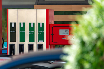 A fuel pump at a gas station displays diesel and super prices on a digital display alongside green foliage. © Anselm