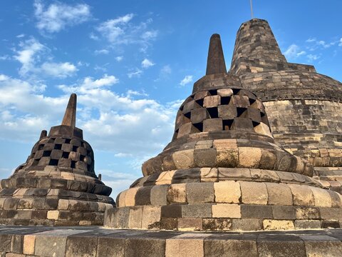 Highly detailed stone stupas at Borobudur Temple, Indonesia; a photo set against a backdrop of a bright blue sky and scattered clouds.