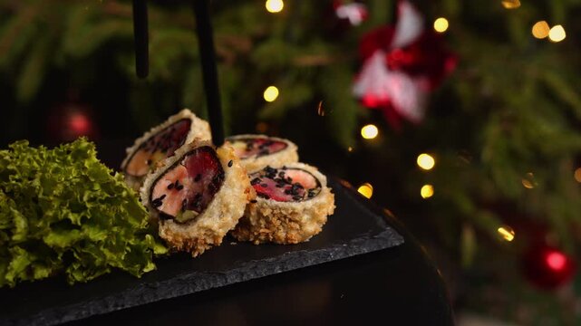 Hand picking tempura sushi roll with chopsticks from slate plate during Christmas dinner, festive holiday lights and pine tree background with bokeh effect and glitter falling