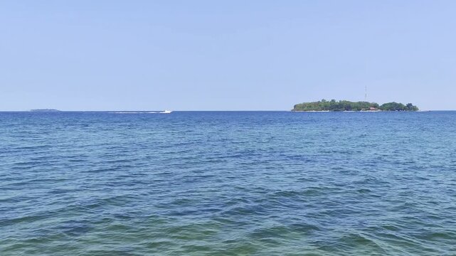 A wide-angle video shot capturing a white motorboat cruising at high speed from left to right across a calm blue sea, creating a white water wake in front of a small green tropical island
