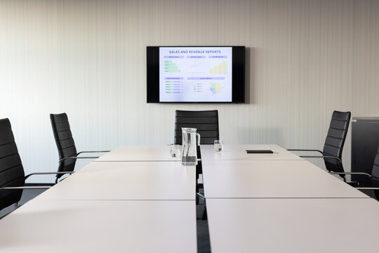 Long white boardroom table extending, black chairs lining sides, monitor displaying sales charts