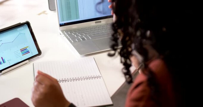 Pen is pointing at laptop spreadsheet with tablet charts on desk, notebook opening to record data
