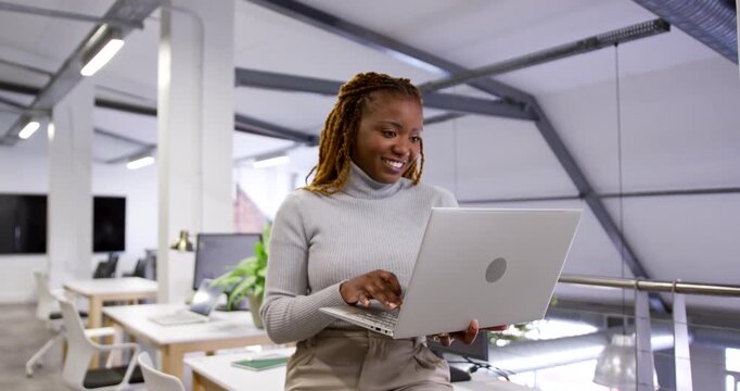 African woman in turtleneck with silver laptop, typing, laughing after ping at mezzanine