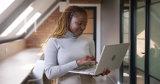 Mid-adult African woman glancing down at silver laptop, typing while wearing gray top in loft hall