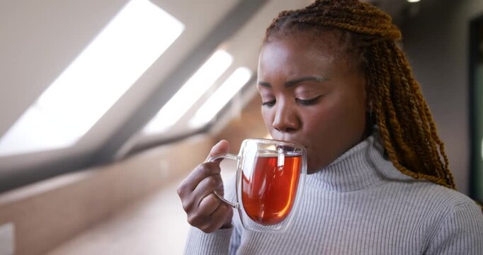 African woman lifting clear mug, sniffing, sipping tea, savoring calm under loft skylights