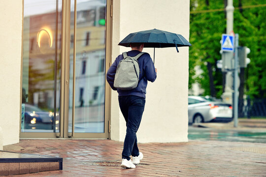 Person walking away on wet brick sidewalk holding open black umbrella wearing blue sweatshirt jeans and gray backpack in rainy urban setting. Pedestrian with umbrella and backpack on rainy city street