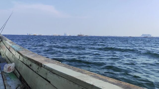 A point-of-view (POV) video shot taken from the side hull of a traditional wooden fishing boat cruising through choppy blue sea water, showcasing gentle waves crashing against the wooden frame under