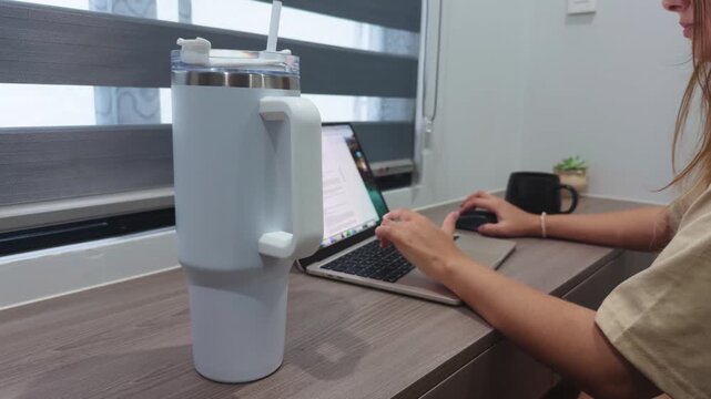 Woman focusing on work, typing on a laptop with an external mouse at a desk in a home office, a large insulated drink container keeping her hydrated and productive during remote working